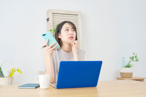パソコンの前でスマートフォンを持って悩む人